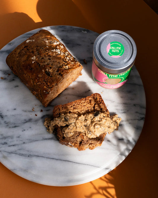 Bananenbrot und ein Glas Full Power Peanut Butter auf einem Marmorteller, der Hintergrund ist orange.