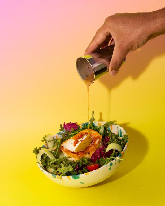 Salatschüssel mit Rucola, Tomaten, Salatgurke und Ziegenkäsetaler, über die ein Peanut Butter Dressing gegossen wird.