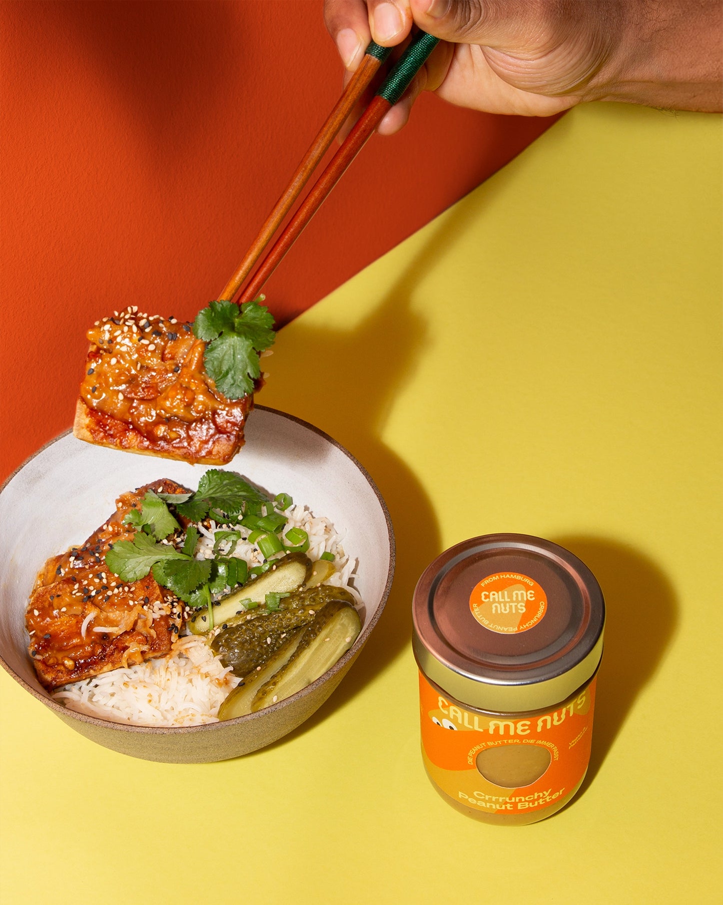 Schüssel mit mariniertem Tofu, Reis und Gewürzgurken und Crrrunchy Peanut Butter Glas vor gelbem Hintergrund
