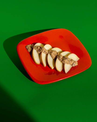 Apfel mit Full Power Peanut Butter auf rotem Teller vor grünem Hintergrund