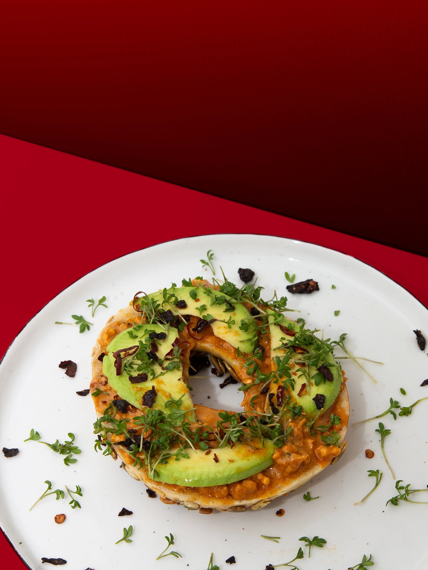 Bagel mit Smokin' Hot Peanut Butter, Avocado und Kresse auf weißem Teller vor rotem Hintergrund