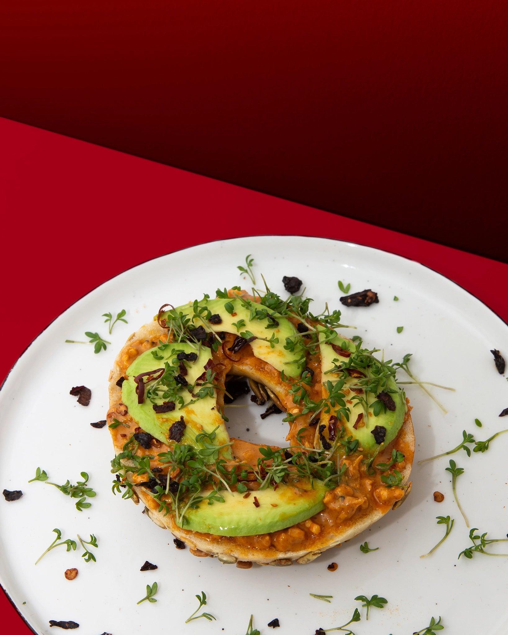 Bagel mit Smokin' Hot Peanut Butter, Avocado und Kresse auf weißem Teller vor rotem Hintergrund
